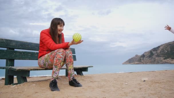 Jeune femme caucasienne avec un bébé sur l'aire de jeux au bord de la mer. Blonde mignon bébé vient à rire maman assis sur le banc pour prendre la balle en caoutchouc jaune avec boutons . — Video