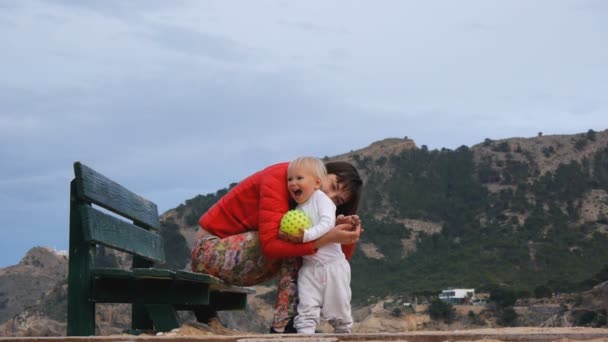 Женщина играет с ребенком на горном фоне. Молодая белая мама, сидящая на скамейке, смешит блондинку. Ребенок держит желтый резиновый мяч с прыщами . — стоковое видео