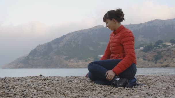 若い海の背景の女性。赤いバブル ジャケットとジーパン海の背景の小石のビーチの上に座って魅力的な白人女性。女の子に触れる手のひらに手のひらから石. — ストック動画