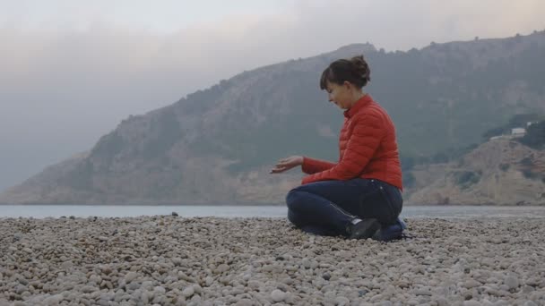 Mladá žena na pozadí moře. Atraktivní Kavkazský žena v červené bublina bunda a džíny sedí na oblázkové pláži na pozadí moře. Dívka spěchá oblázky z její dlaně. — Stock video