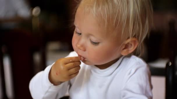 Bébé manger de la nourriture au restaurant. Blonde enfant aux yeux bleus en chaise haute prenant un morceau de nourriture à la main à partir d'une assiette le mettant dans la bouche . — Video