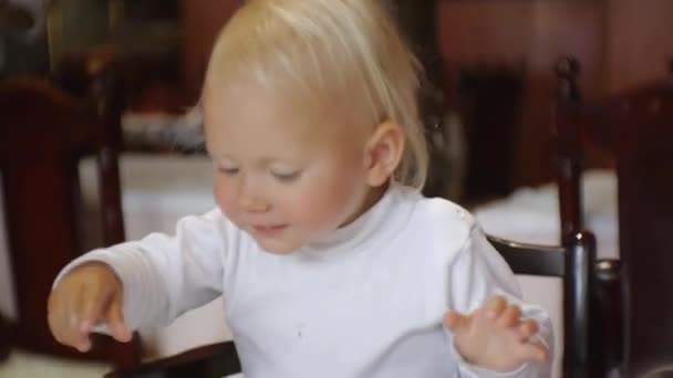 Bebé comiendo comida en el restaurante. Feliz niño de ojos azules en silla alta tomando pedazo de pasta a mano de un plato poniéndola en la boca . — Vídeo de stock