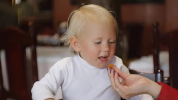 Baby eten in restaurant. Blonde blauwogige kind in de kinderstoel nemen van voedsel door de mond met de moeders handen. — Stockvideo