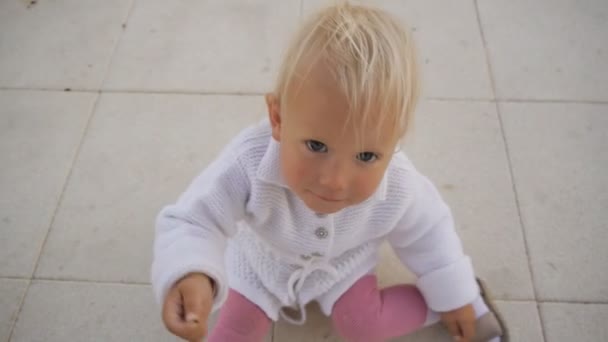 ニットの白のドレスで金髪の青い目の赤ちゃん女の子は笑って彼女の手で小さな蟻を示す地面に座っています。上記からの眺めを撮影しました。幸せな子供時代の概念. — ストック動画
