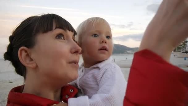 Donna attraente con bambino sul lungomare di sera. Primo piano di mamma che mostra la città alla bambina tra le braccia sullo sfondo della costa del mare. Concetto di infanzia felice e famiglia . — Video Stock