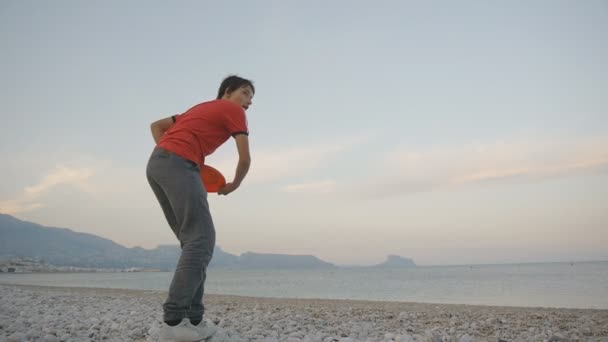 Adolescente tocando disco volador. Bajo ángulo de tiro de caucásico adolescente lanzando disco volador en una playa de guijarros en el fondo de la costa de montaña . — Vídeos de Stock