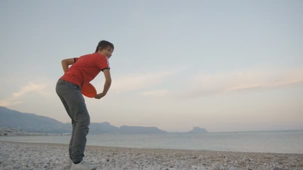 Підліток грає на літаючому диску. Низький кут пострілу радісного хлопчика-підлітка кидає літаючий диск на гальковому пляжі на фоні гірського узбережжя . — стокове відео
