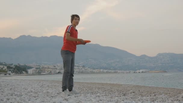 Adolescent jouant un disque volant. Caucasien adolescent garçon jeter disque volant sur une plage vide basse saison sur fond de bord de mer de montagne . — Video