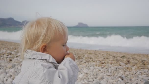 Portrait d'une petite fille blonde caucasienne mangeant un fruit d'un médaillon avec des larmes aux yeux sur fond de vagues de mer orageuses . — Video