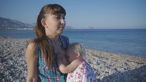 年轻的现代白种母亲母乳喂养她的金发女孩在空荡荡的日落海滩上. — 图库视频影像