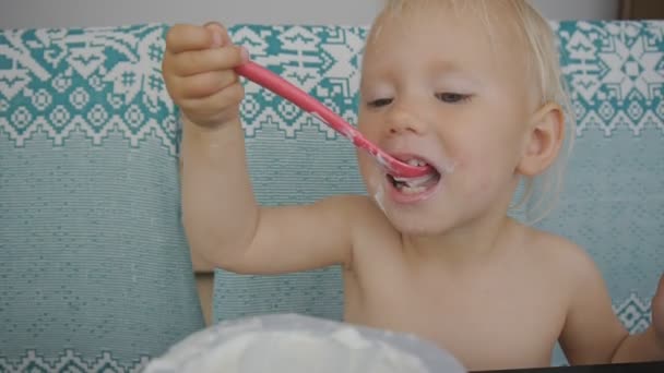 Bebé comiendo yogur haciendo un desastre. Primer plano de linda niña de ojos azules degustación de yogur con una mueca . — Vídeos de Stock