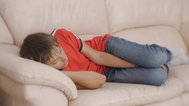 Tonåringen sover på soffan. Kaukasiska teen pojke i röd t-shirt och blå jeans sover på beige skinnsoffa i dagtid. Brist på sömn. — Stockvideo