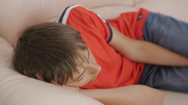 Teenager sleeping on couch. Caucasian teen boy in red t-shirt and blue jeans sleeping on beige leather sofa in daytime. Lack of sleep. — Stock Video