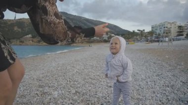 Genç Anne ile bebek günbatımı sahilde. Siyah dar elbiseli çekici beyaz kadın boş çakıl plaj zemin üzerine iki yaşında çocuk yetiştirme eller uzanır. Mutlu aile kavramı.