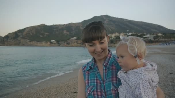 Smiling attractive caucasian woman in plaid blue shirt with baby girl with glasses for swimming walking on pebble beach along the sea. — Stock Video