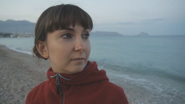 Jonge vrouw op lege strand achtergrond. Close-up van aantrekkelijke Kaukasische bruin donkerharige vrouw in Rode hoodie kijken naar de zee. — Stockvideo