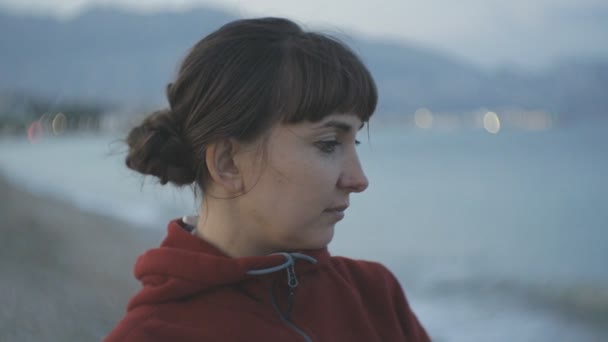 Young woman on empty beach background. Portrait of attractive caucasian brown haired female in red hoodie looking at the sea. — Stock Video