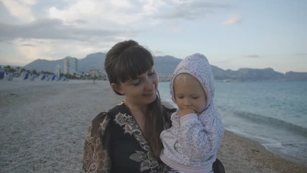 Genç Anne ile bebek günbatımı sahilde. Siyah dar elbiseli iki yaşında çocuk kucağında ile boş çakıl plaj arka plan üzerinde yürüme çekici beyaz kadın. Mutlu aile kavramı. — Stok video