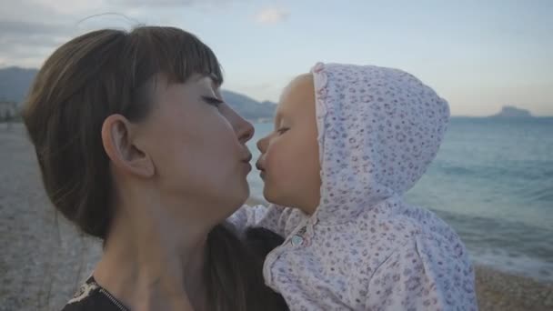 Ung mamma med bebis på solnedgång stranden. Närbild av attraktiv kaukasisk kvinna kysser två år gamla barn på tom pebble beach bakgrund. Begreppet lycklig familj. — Stockvideo