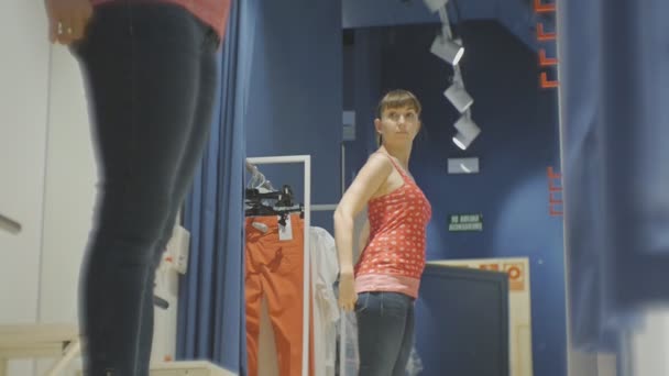 Jonge vrouw proberen op kleding. Lage hoekmening van aantrekkelijke Kaukasische vrouwelijke kijken in de spiegel spinnen proberen op rode shirt en spijkerbroek in kleding winkels paskamer. — Stockvideo