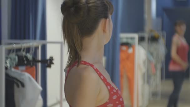 Fiatal nő megpróbálja a ruhákat. Vonzó kaukázusi nő néz a tükörbe, spinning körül kipróbálás-ra piros inget a próbafülke ruházati üzletek. — Stock videók