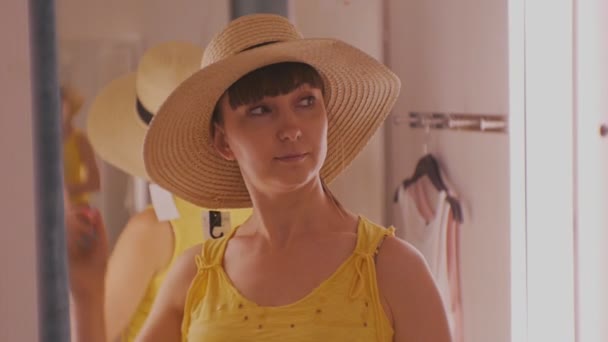 Young woman trying on clothes. Attractive caucasian female looks in the mirror turning her head trying on broad-brimmed straw hat in clothing stores fitting room. — Stock Video