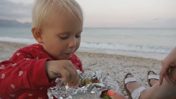 Close-up van twee jaar oude baby eten van een lepel met voedsel uit de lade van een folie op het strand. Kind en moeder op de zonsondergang strand. — Stockvideo