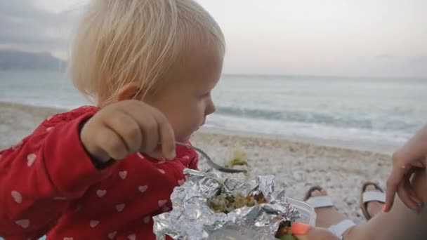 Close-up van twee jaar oude baby eten van een lepel met voedsel uit de lade van een folie op het strand. Kind en moeder op de zonsondergang strand. — Stockvideo