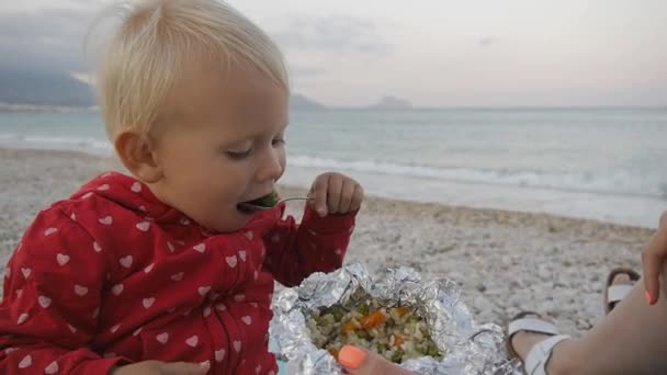 Porträt eines zweijährigen Babys, das grüne Oliven aus einem Folientablett am Strand isst. Kind und Mutter am Strand des Sonnenuntergangs. — Stockvideo