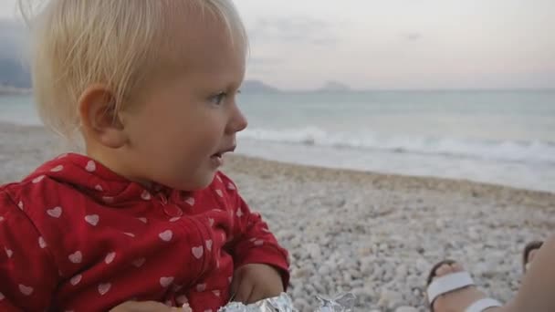 Portrait of two years old baby chewing food on the seaside background. Child and mom on the sunset beach. — Stock Video