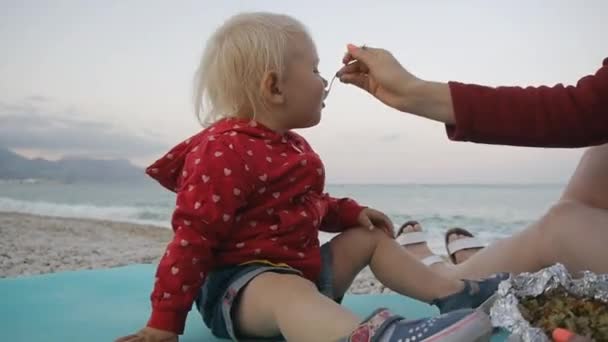 Blond meisje plezier eten van een lepel met voedsel uit de hand van een moeders op de achtergrond van de zee. Kind en moeder op de zonsondergang strand. — Stockvideo