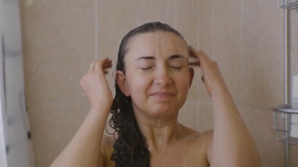 Uma mulher a tomar banho. Retrato de jovem caucasiana com os olhos fechados de pé sob o chuveiro enxaguando o cabelo com água . — Vídeo de Stock