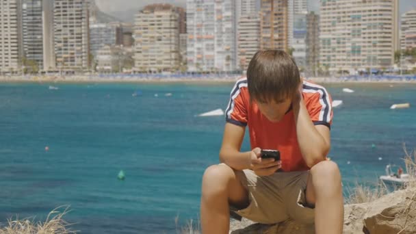 Caucasico teen ragazzo si siede su pietra guardando smartphone schermo sul mare costa grattacieli skyline sfondo . — Video Stock