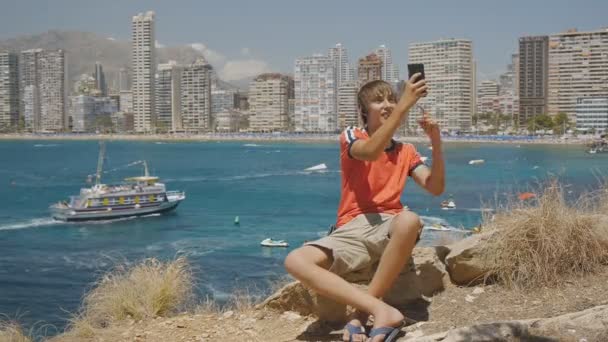 Caucásico adolescente chico hace videollamada en el teléfono inteligente que muestra lugares de interés en la costa del mar y rascacielos skyline fondo . — Vídeos de Stock