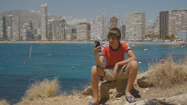 Stilig kaukasiska teen pojke ler vilket gör smartphone video kalla på havet kusten och skyskrapor skyline bakgrund. — Stockvideo