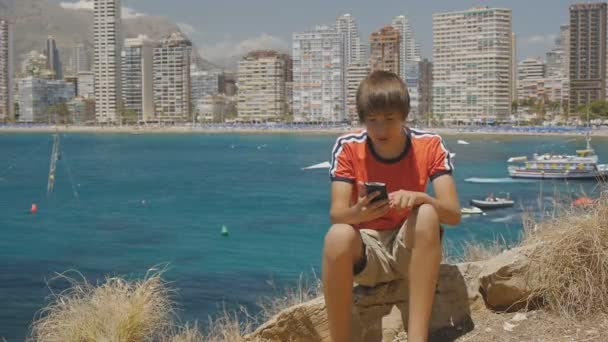 15 år gammal teen pojke typer meddelande på smartphone som sitter på stenen på havet kusten och skyskrapor skyline bakgrund. — Stockvideo