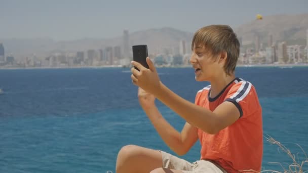 Retrato de adolescente guapo en camiseta roja y pantalones cortos haciendo videollamada de teléfonos inteligentes en el fondo del horizonte de la ciudad de la costa del mar . — Vídeos de Stock