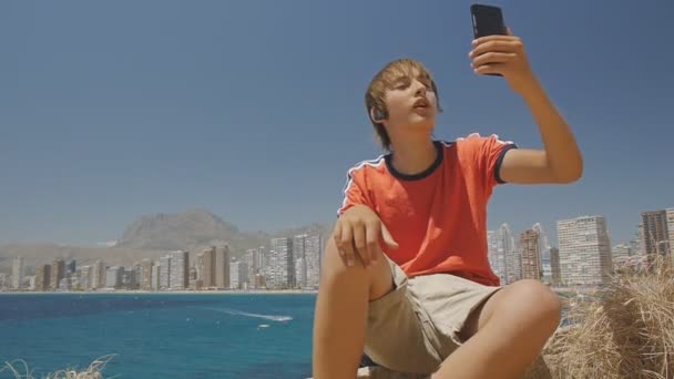 Милий хлопчик робить відеодзвінок на смартфон, показуючи приціли навколо на морському узбережжі горизонту міста фону . — стокове відео