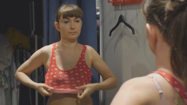 Jonge vrouw proberen op kleding. Bruin-haired Kaukasische vrouw in een beige beha kijkt in de spiegel rood shirt in kledingwinkels montage kamer zetten. — Stockvideo
