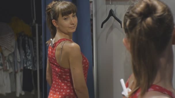 Νεαρή γυναίκα που προσπαθούν για ρούχα. Ελκυστική καυκάσιος γυναίκα κοιτάζει στον καθρέφτη περιστρέφεται γύρω από την προσπάθεια στο κόκκινο πουκάμισο σε καταστήματα ειδών ένδυσης δοκιμαστήρια. — Αρχείο Βίντεο