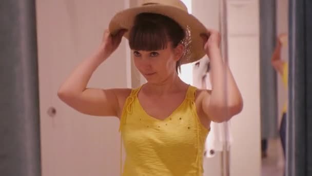 Young woman trying on clothes. Smiling caucasian female looks in the mirror turning her head trying on broad-brimmed straw hat in clothing stores fitting room. — Stock Video