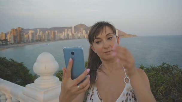 Attraktiv ung kvinna tar selfie med smartphone. Kaukasiska tjej att göra självporträtt foto eller video med mobiltelefon på sommar solnedgång seaside resort city bakgrund. — Stockvideo