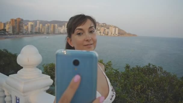 Vonzó fiatal nő vesz selfie smartphone. Mosolyogva kaukázusi nő így önarckép fénykép vagy mozgókép mellékelése mobiltelefon nyári naplemente seaside resort city háttér. — Stock videók