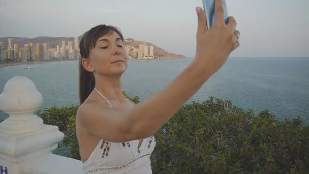 Jolie jeune femme prenant selfie avec smartphone. Jolie fille souriante faisant photo autoportrait ou vidéo avec téléphone portable sur fond de ville station balnéaire coucher de soleil d'été . — Video