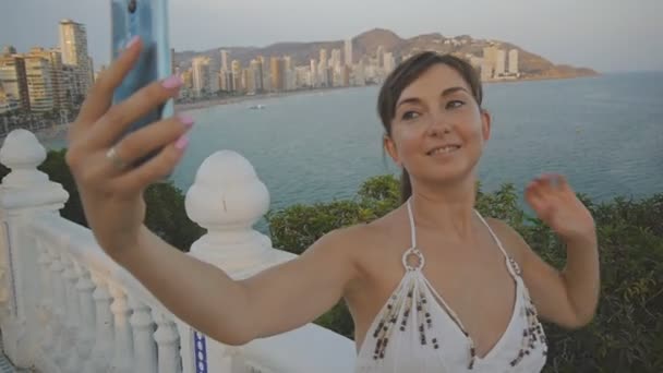 Çekici genç kadın selfie smartphone ile alıyor. Kendi kendine portre fotoğraf veya video cep telefonu ile yaz günbatımı sahil resort şehir arka plan üzerinde yapma güzel gülümseyen kız. — Stok video