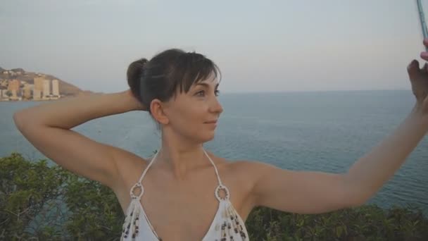 Attraktiv ung kvinna tar selfie med smartphone. Söt tjej att göra självporträtt foto eller video med mobiltelefon röra hennes hår på sommaren sunset seaside resort city bakgrund. — Stockvideo