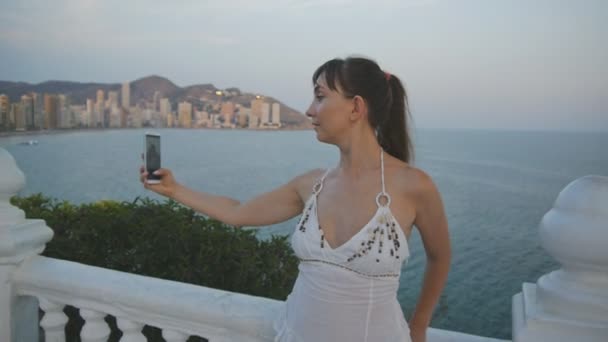 Привлекательная молодая женщина делает селфи со смартфоном. Улыбающаяся девушка делает автопортретное фото или видео с мобильным телефоном на летнем закате на приподнятом балконе . — стоковое видео
