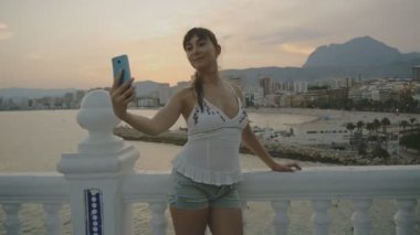Smartphone ile çekici genç kadın alarak selfie. Seksi beyaz kadın kendi kendine portre fotoğraf veya video cep telefonu ile yaz sunset resort Şehir dağ ve deniz manzara arka plan üzerinde yapma.