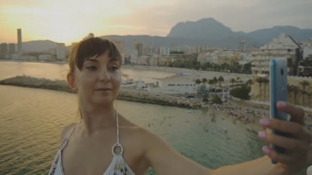 Привлекательная молодая женщина делает селфи со смартфоном. Улыбающаяся кавказская женщина делает автопортрет фото или видео с сотовым телефоном на летнем курорте закат города горы и морской пейзаж фоне . — стоковое видео