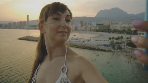 Atrakcyjnych młodych kobiet biorąc selfie z smartphone. Uśmiechający się kaukaski kobieta co autoportret zdjęcie lub nagranie wideo z telefonu komórkowego na lato: sunset resort miasta góry i morze krajobraz tła. — Wideo stockowe
